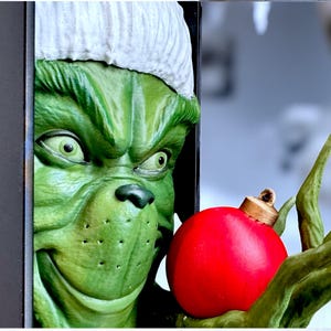 May include: A green, cartoonish character with a white hat and a mischievous grin, holding a bright red Christmas ornament. The character's face is detailed with green skin, and the ornament is round with a gold cap.