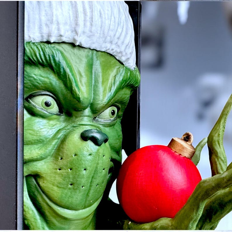 May include: A green, cartoonish character with a white hat and a mischievous grin, holding a bright red Christmas ornament. The character's face is detailed with green skin, and the ornament is round with a gold cap.