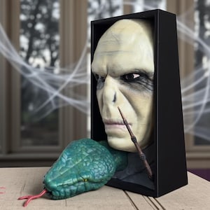 May include: A sculpture of a pale-skinned face with a wand, emerging from a black box. A green snake with a red tongue is positioned in front of the box. The sculpture has a detailed, realistic appearance.