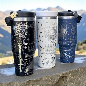 May include: Three insulated travel mugs with detailed silver designs. One is black with text and a sword, another is white with a mountain scene, and the third is navy blue with celestial motifs. Each mug has a black or white lid with a handle.