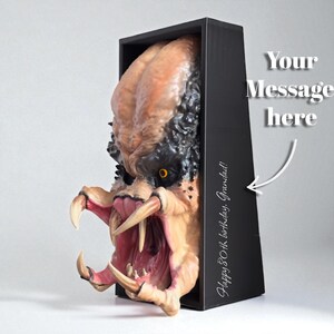 May include: A Predator mask sculpture, featuring detailed skin textures and sharp teeth, emerges from a black box. The mask is painted in shades of tan, black, and pink, with yellow eyes. The box has the text "Happy 80th birthday, Grandad!" and "Your Message here".