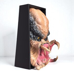 May include: A detailed sculpture of a Predator alien head, featuring textured skin tones of tan and black, with sharp teeth and a gaping mouth. The sculpture is set within a black rectangular frame, creating a 3D effect.