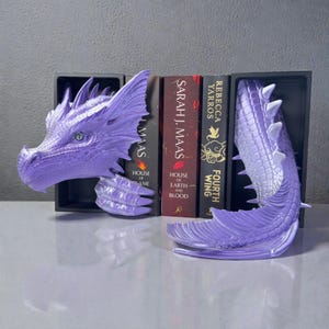 May include: A pair of lavender dragon bookends. The dragon head and tail are detailed with scales and spikes. The bookends are holding up several books with titles including "House of Earth and Blood" and "Fourth Wing".