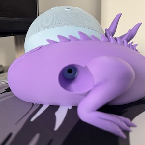 Pode incluir: Um suporte de alto-falante inteligente em forma de lagarto, impresso em 3D, de cor roxa, com um furo na parte de tr&aacute;s para segurar um pequeno alto-falante inteligente azul claro.