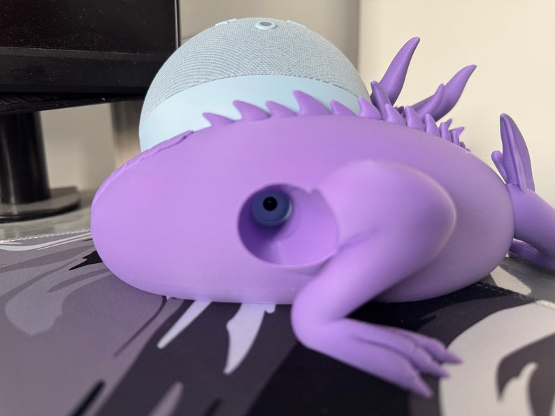 Pode incluir: Um suporte de alto-falante inteligente em forma de lagarto, impresso em 3D, de cor roxa, com um furo na parte de tr&aacute;s para segurar um pequeno alto-falante inteligente azul claro.