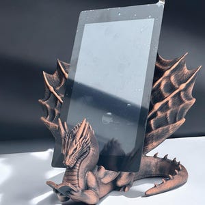 May include: A bronze dragon figurine holding a tablet in its claws. The dragon has a detailed design with wings and a long tail. The tablet is black and has a screen protector.