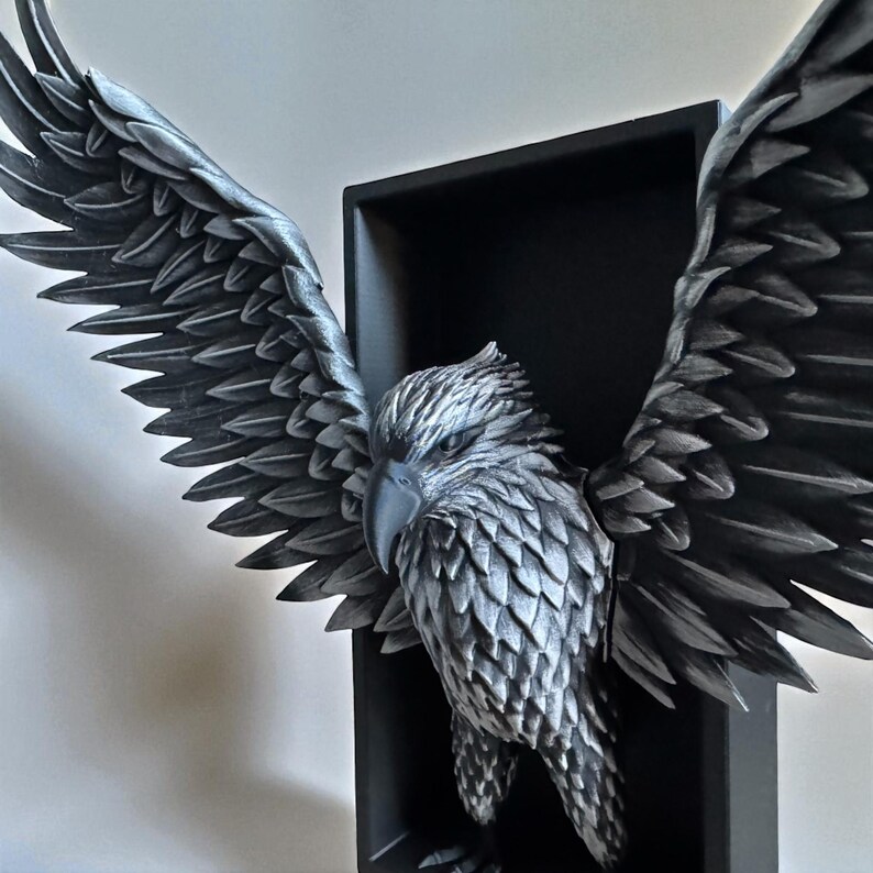 May include: A detailed, grayscale sculpture of an eagle with outstretched wings, mounted within a black rectangular frame. The eagle's feathers are intricately detailed, and the sculpture is set against a white wall.