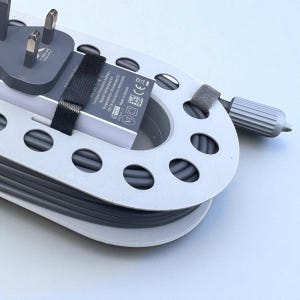 May include: A gray power cord neatly coiled within a white and gray cable organizer. The organizer has a series of oval cutouts and a small gray handle. A gray power adapter with three prongs is visible.