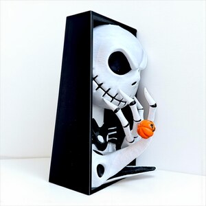 May include: A white and black sculpture of a character with a skeleton face and a black cloak. The character is holding a small orange pumpkin. The sculpture is in a black frame.