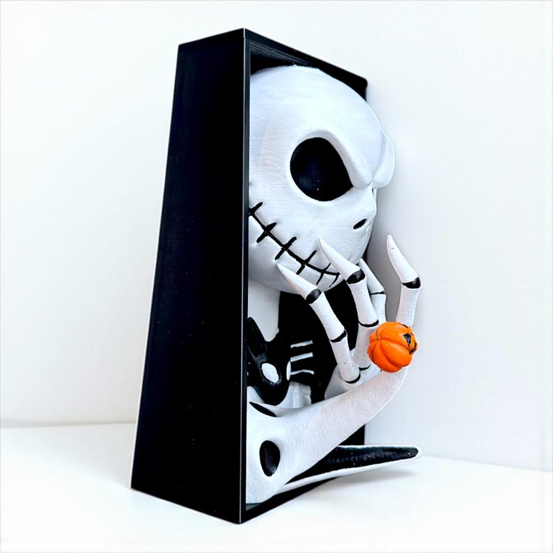 May include: A white and black sculpture of a character with a skeleton face and a black cloak. The character is holding a small orange pumpkin. The sculpture is in a black frame.