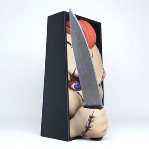 Puede incluir: Una caja decorativa con tem&aacute;tica de Chucky que presenta una cabeza de mu&ntilde;eco Chucky con cabello naranja, ojos azules y detalles faciales cosidos. La mu&ntilde;eca sostiene un gran cuchillo plateado. La caja es negra y tiene forma rectangular.