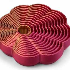 May include: A 3D-printed, flower-shaped object in shades of red and gold. The design features concentric, wavy layers, creating a textured, layered appearance. The centre has a golden hue, contrasting with the red outer layers. The object is on a white background.