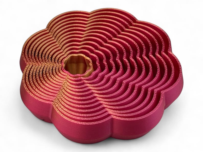 May include: A 3D-printed, flower-shaped object in shades of red and gold. The design features concentric, wavy layers, creating a textured, layered appearance. The centre has a golden hue, contrasting with the red outer layers. The object is on a white background.