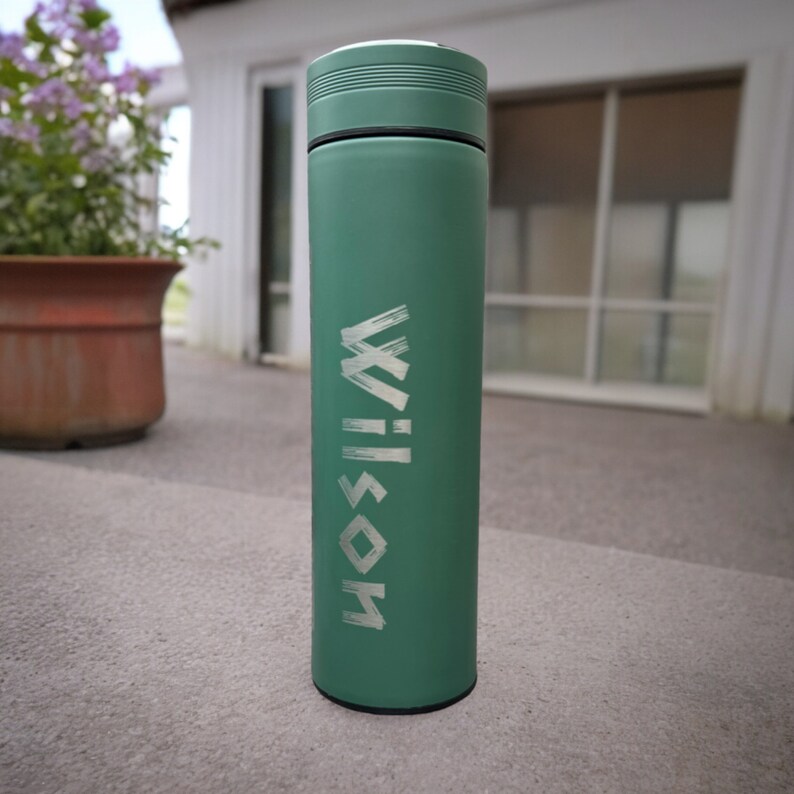 A tumbler standing on a concrete surface outside a house. The tumbler features the name Wilson written. In the background, there is a window with closed blinds and a potted plant with purple flowers.