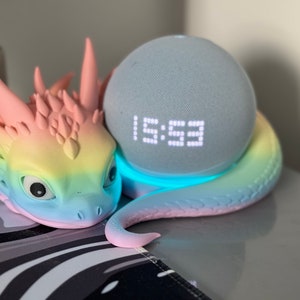Pode incluir: Uma figura de drag&atilde;o com cores de arco-&iacute;ris com um alto-falante inteligente azul claro aninhado em suas espirais. O rel&oacute;gio digital do alto-falante mostra a hora 1:59.