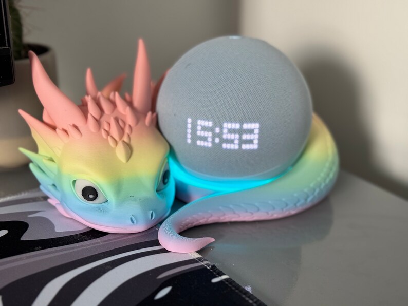 Pode incluir: Uma figura de drag&atilde;o com cores de arco-&iacute;ris com um alto-falante inteligente azul claro aninhado em suas espirais. O rel&oacute;gio digital do alto-falante mostra a hora 1:59.
