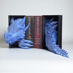 May include: Blue dragon bookends with a detailed, textured design. The dragon head and tail are positioned on either side of the books. The books are titled "House of Earth and Blood" and "House of Sky and Shadow" by Sarah J. Maas.
