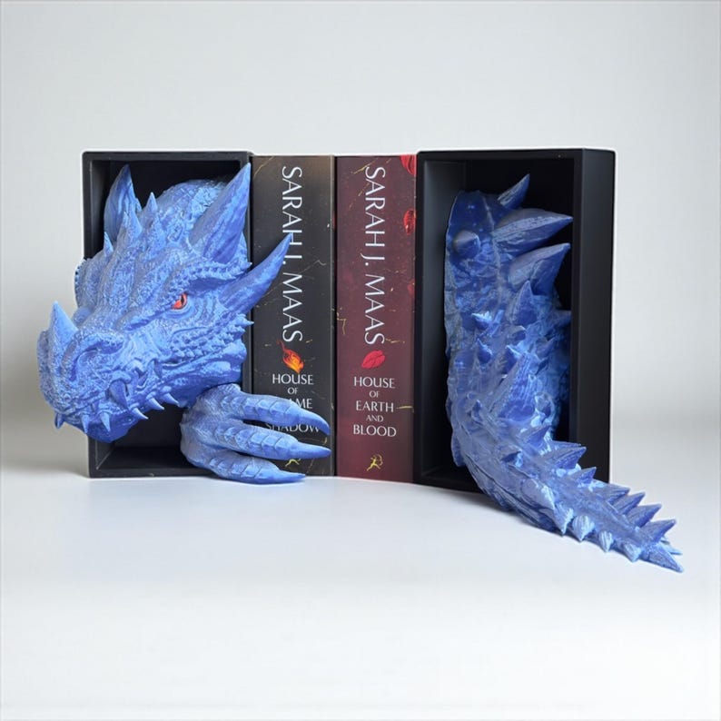 May include: Blue dragon bookends with a detailed, textured design. The dragon head and tail are positioned on either side of the books. The books are titled "House of Earth and Blood" and "House of Sky and Shadow" by Sarah J. Maas.