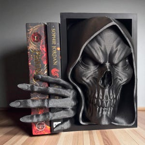 May include: A black, 3D-printed bookend depicting a skull in a hooded cloak and a skeletal hand holding two books. The books have red and gold covers, with the title "PHANTAS" visible. The bookend is on a wooden surface.