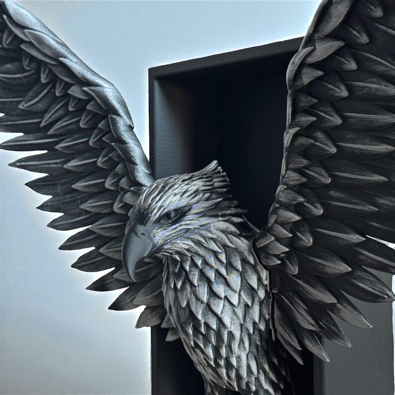 May include: A detailed, metallic gray eagle sculpture with outstretched wings, mounted within a black rectangular frame. The eagle's feathers and facial features are intricately carved, creating a textured appearance. The sculpture is a decorative piece.