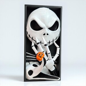 May include: A white and black framed sculpture of Jack Skellington from The Nightmare Before Christmas. The sculpture features Jack's skeletal face, hands, and a small orange pumpkin. The background is black.