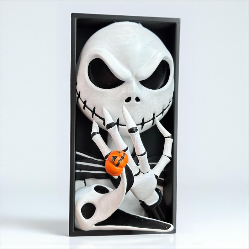 May include: A white and black framed sculpture of Jack Skellington from The Nightmare Before Christmas. The sculpture features Jack's skeletal face, hands, and a small orange pumpkin. The background is black.
