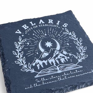 Peut inclure: Un dessous de verre en ardoise noire avec un motif blanc repr&eacute;sentant une lune croissante, une &eacute;toile, des montagnes et un livre ouvert. Le texte "VELARIS CITY OF STARLIGHT" et "To the stars who listen and the dreams that are answered" est &eacute;galement inclus dans le motif.