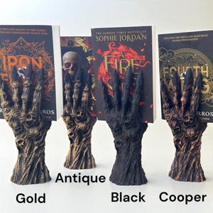 May include: Four decorative hand-shaped bookends in gold, antique, black, and copper. Each bookend holds a book with titles like "Iron Flame" and "A Fire Within". The bookends have a detailed, textured design and are labeled with their respective colors.