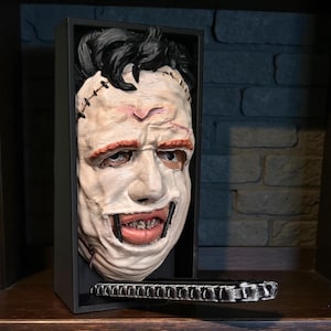 May include: A detailed, three-dimensional mask of a character from a horror film, displayed in a black box. The mask is white with red accents around the eyes and mouth, and black hair. The mouth is open, revealing teeth. The box has a black base.