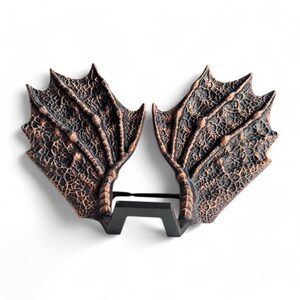 May include: A pair of dragon wings made of a textured, dark brown material with a copper finish. The wings are attached to a black metal stand.