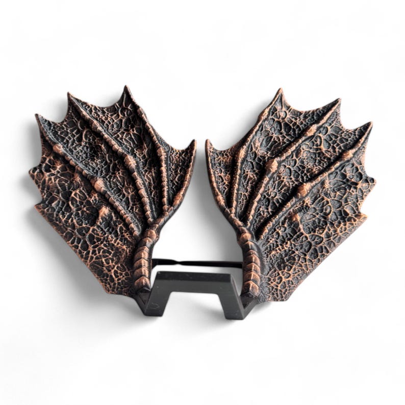 May include: A pair of dragon wings made of a textured, dark brown material with a copper finish. The wings are attached to a black metal stand.