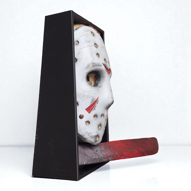 May include: A white hockey mask with red accents and a black frame. The mask has multiple holes and red slashes. A red, blood-stained machete extends from the bottom of the mask. The item is a decorative piece.