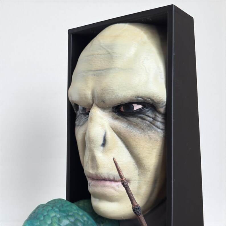 May include: A detailed sculpture of a character's face, framed in black. The face is pale with dark eye sockets and a prominent nose. A wand is held near the mouth. The sculpture is likely a collectible or decorative item.