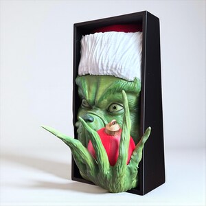 May include: A green Grinch figure wearing a Santa hat, holding a red Christmas ornament. The Grinch's face is detailed with a grumpy expression. The figure is set in a black rectangular frame, perfect for holiday decor.