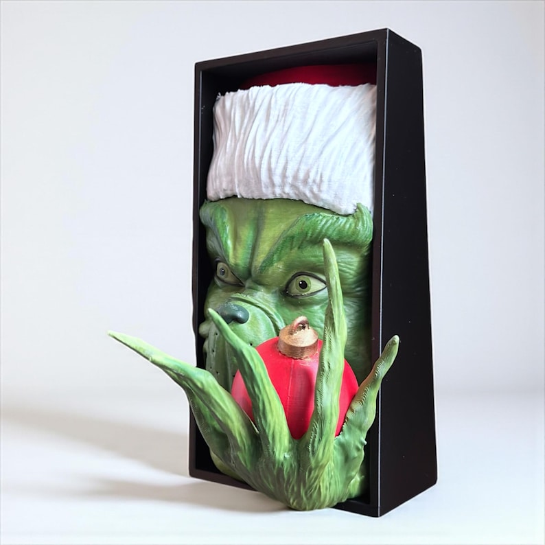 May include: A green Grinch figure wearing a Santa hat, holding a red Christmas ornament. The Grinch's face is detailed with a grumpy expression. The figure is set in a black rectangular frame, perfect for holiday decor.