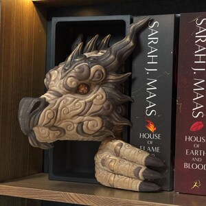 May include: A detailed dragon head and claw bookend in shades of beige, brown, and gold. The dragon's head features intricate patterns and an orange eye. The bookend is designed to hold books, with the title "House of Flame" visible.