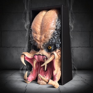May include: A detailed sculpture of a Predator alien head, featuring a textured, tan and black exterior with yellow eyes and sharp teeth. The sculpture is set within a black rectangular frame, creating a 3D effect.