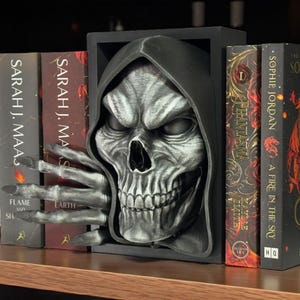May include: A detailed, black and grey skull-shaped bookend with a skeletal hand extending outwards. The bookend is positioned between books, including titles such as "Flame and Shadow" and "A Fire in the Sky". The skull has a textured finish.