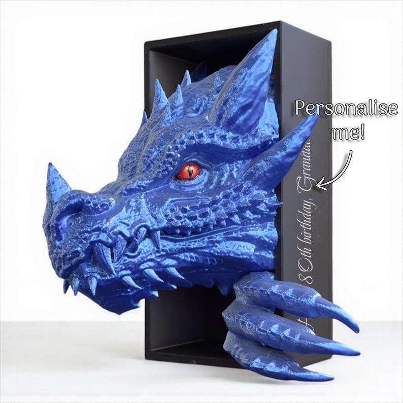 May include: A blue dragon head sculpture with red eyes and sharp teeth emerges from a black box. The dragon's head is highly detailed, with textured scales and horns. Text on the side says "Personalise me!" and "80th birthday, Grandma".