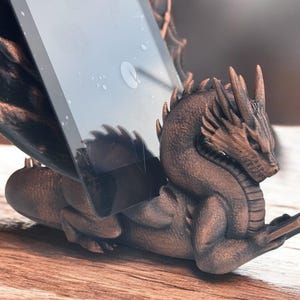 May include: A bronze dragon phone stand with a tablet propped up on its back. The dragon is holding a book in its front paws.
