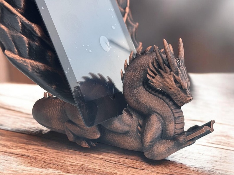 May include: A bronze dragon phone stand with a tablet propped up on its back. The dragon is holding a book in its front paws.
