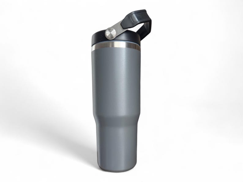 May include: A grey insulated tumbler with a black lid and handle. The stainless steel rim and handle hardware provide a metallic accent. The tumbler is designed to keep beverages cold or hot. The handle is attached to the lid with a hinge.