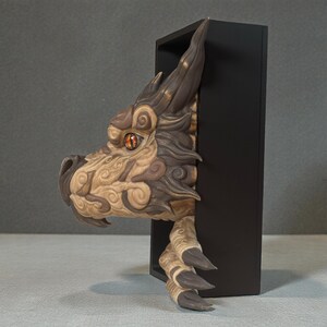 May include: A detailed sculpture of a dragon's head and claw emerging from a black rectangular frame. The dragon features intricate carvings, a brown and tan color scheme, and an orange eye. The claw and head are the focal points of this unique art piece.