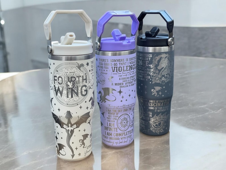 May include: Three stainless steel travel mugs with different designs. The first mug is white with a silver dragon and the text "Fourth Wing". The second mug is lavender with a silver dragon and text about dragons and riders. The third mug is gray with a silver dragon and the text "Only if you are fascinated by the power of this dragon will it without you".