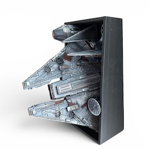 Może przedstawiać: Szczeg&oacute;łowy model statku kosmicznego Millennium Falcon, w odcieniach szarości i brązu, jest prezentowany w czarnej prostokątnej ramie. Model prezentuje skomplikowane panele i szczeg&oacute;ły mechaniczne, z widoczną okrągłą anteną radarową.