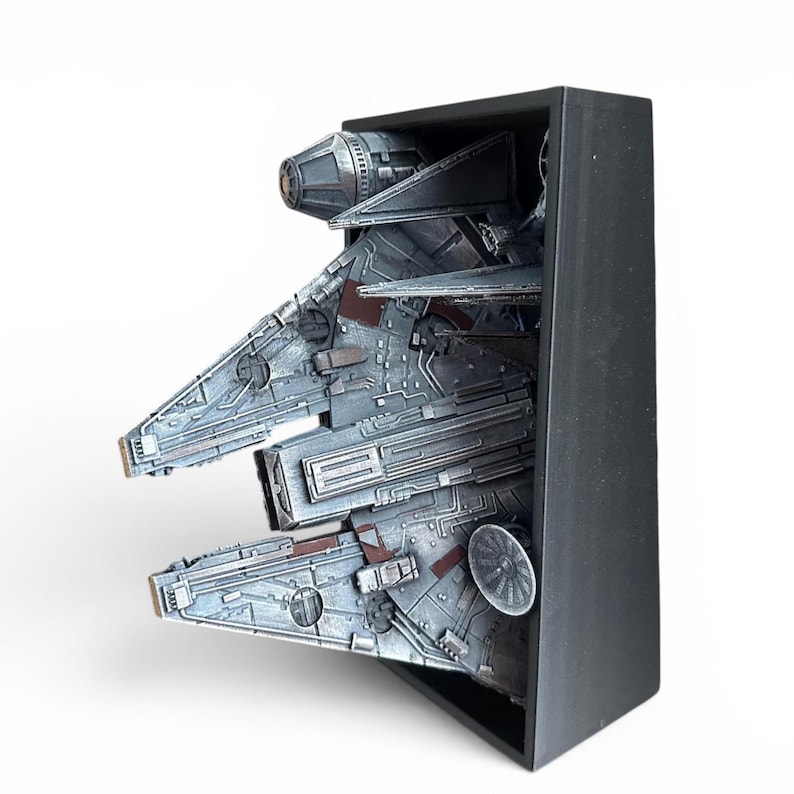 Może przedstawiać: Szczeg&oacute;łowy model statku kosmicznego Millennium Falcon, w odcieniach szarości i brązu, jest prezentowany w czarnej prostokątnej ramie. Model prezentuje skomplikowane panele i szczeg&oacute;ły mechaniczne, z widoczną okrągłą anteną radarową.