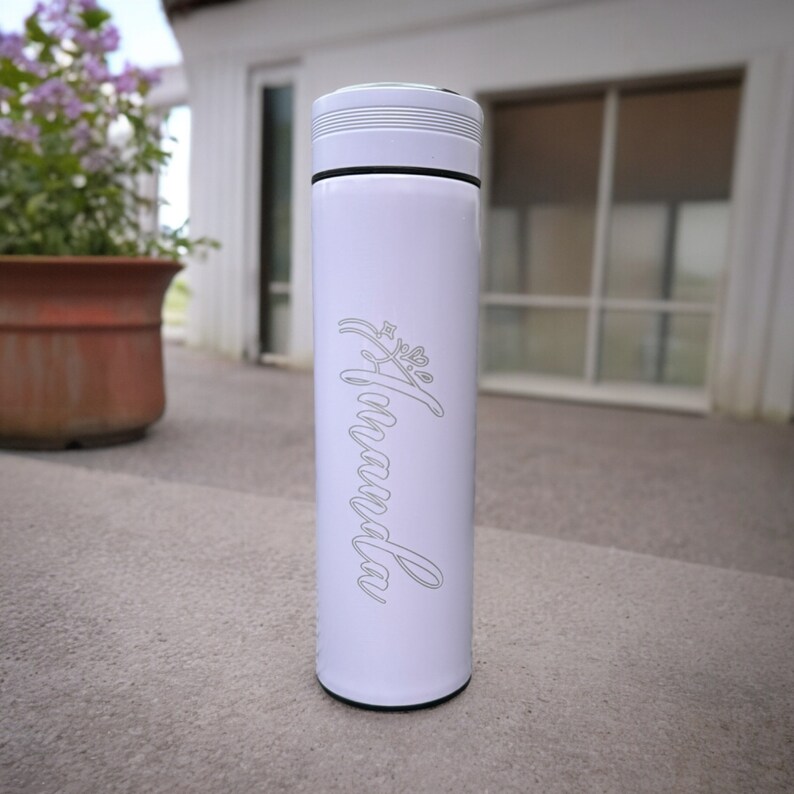 A tumbler standing on a concrete surface outside a house. The tumbler features the name Amanda written in a cursive font with a small decorative flourish. In the background, there is a window with closed blinds and a potted plant with purple flowers.
