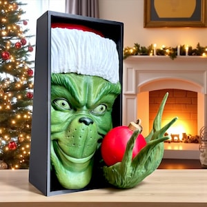 May include: A green character with a Santa hat, holding a red Christmas ornament. The character's face is framed in a black box. The ornament is held in a green hand. Christmas decor.
