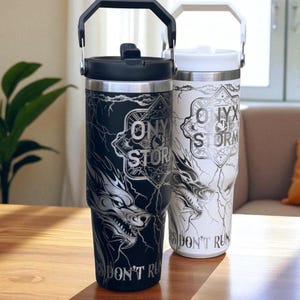 May include: Two insulated travel mugs, one black and one white, with black and white handles. Each mug features a wolf design with the words "ONYX STORM" and "DON'T RUN". The mugs are on a wooden table.
