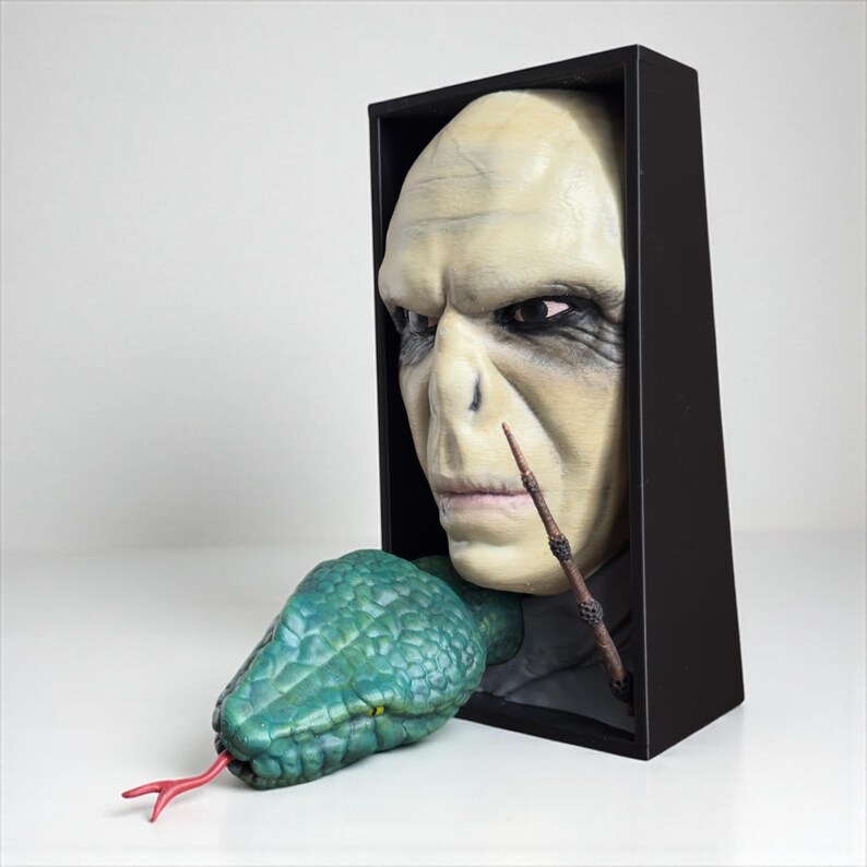 May include: A sculpture featuring a pale-skinned face with a wand and a green snake emerging from a black box. The snake has a red tongue. The face has dark eye makeup and a stern expression.